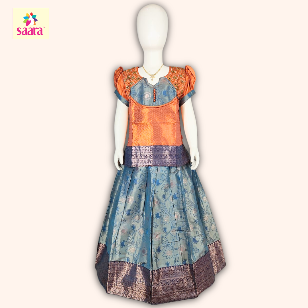 This Pattu Pavadai features a Orange blouse with Aari work, puffed sleeves and a matching Blue skirt made of floral stem designes jacquard fabric.