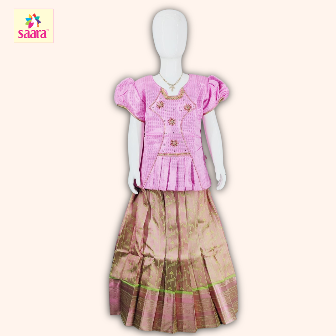 This Pattu pavadai features a Pink blouse with Stone work,Frills,Piping work, puffed sleeves with a matching Copper skirt made of jacquard fabric.